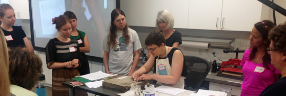 A group of people gathered around a table watching a demonstration by Karen Hanmer working on a bookbinding or craft project in a studio or classroom setting
