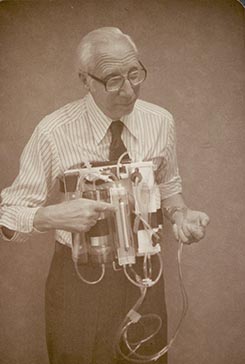 An elderly man in a striped shirt holds a complex, retro-looking device with tubes, wires, and containers attached.