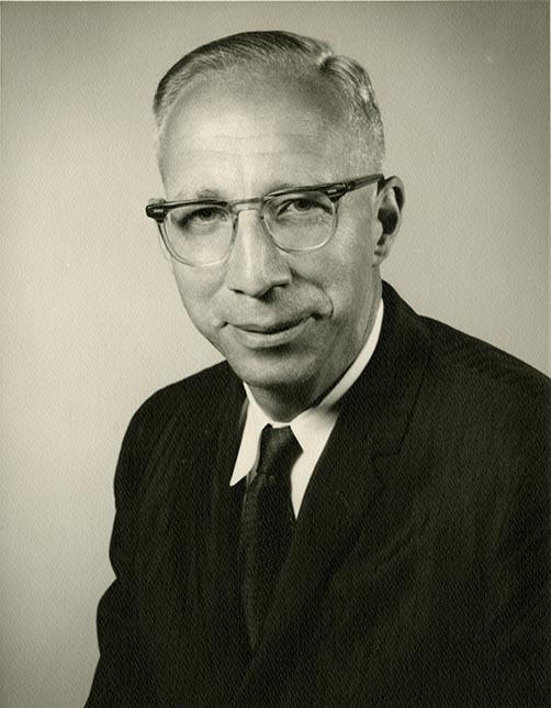 A black-and-white portrait of a man in a formal suit and tie, with neatly combed hair, against a plain background.