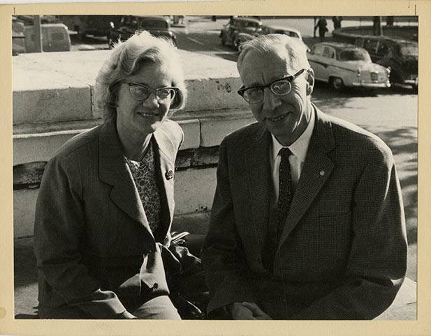 A vintage black-and-white photo of a couple sitting together on a stone wall, with classic cars visible in the background.