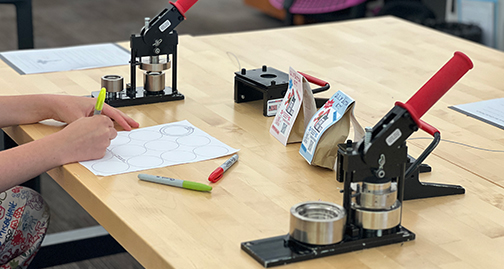 2 button makers on a woodblock table with hands of person coloring a button insert