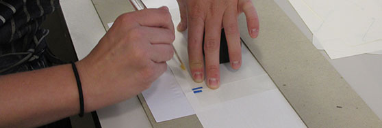 A person using a craft knife to cut or score paper on a work surface, demonstrating bookbinding or similar handcraft techniques