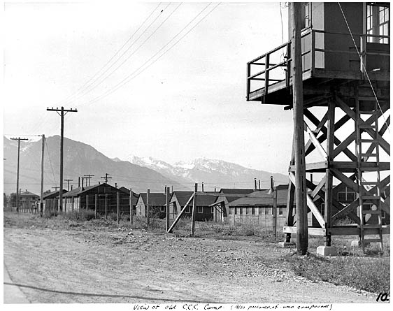 Black-and-white photo of a CCC camp, featuring wooden buildings, a watchtower, and distant mountains under a clear sky.