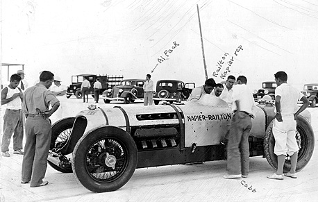 Several men stand around an old race car, engaged in conversation