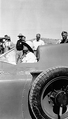 A black and white image of a man inside a race car, surrounded by other people in the Salt Flats of Utah, showcasing the sleek design of the vehicle