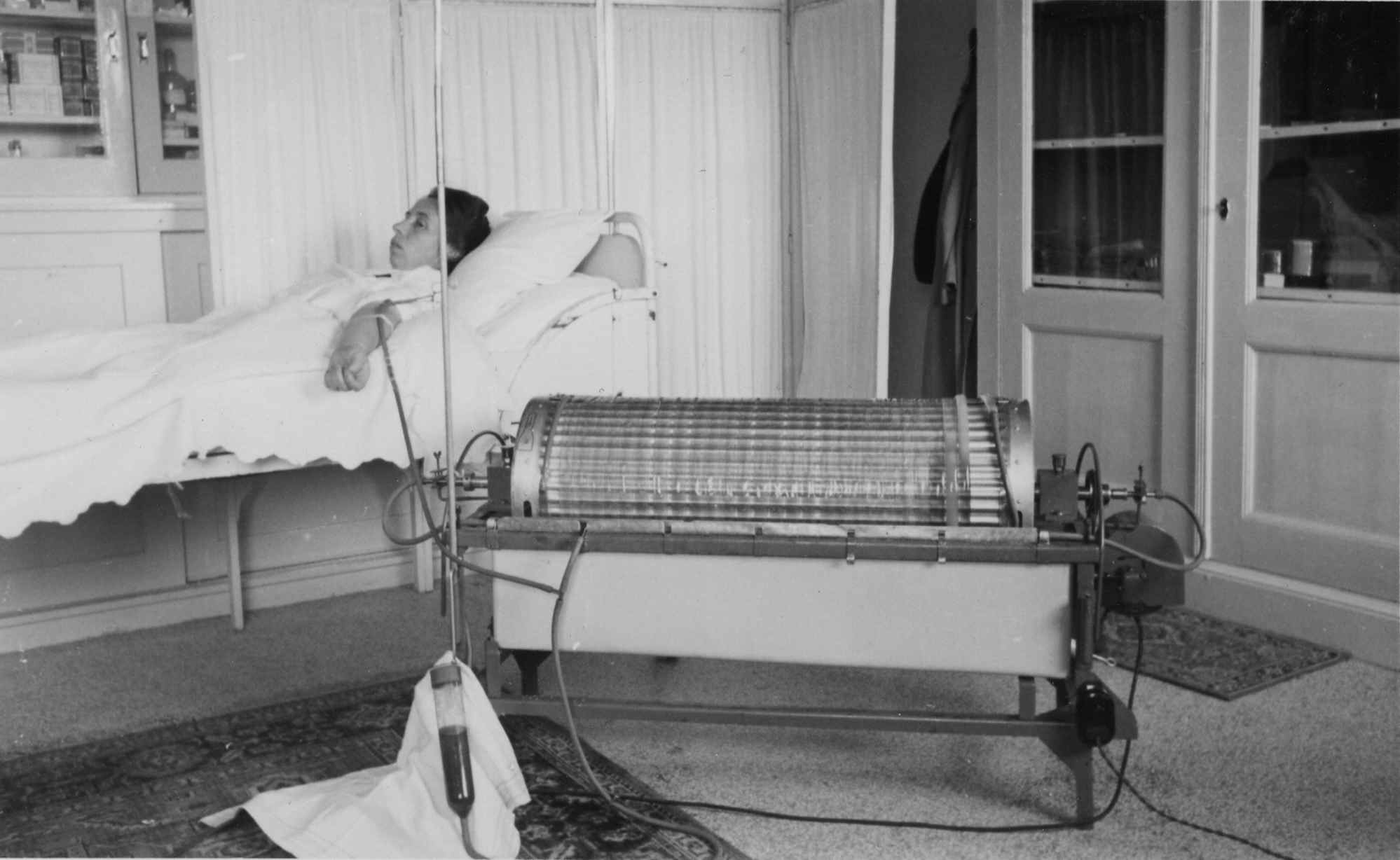 A hospital room featuring a patient on a bed next to an antique medical device with a cylindrical apparatus and control knobs.