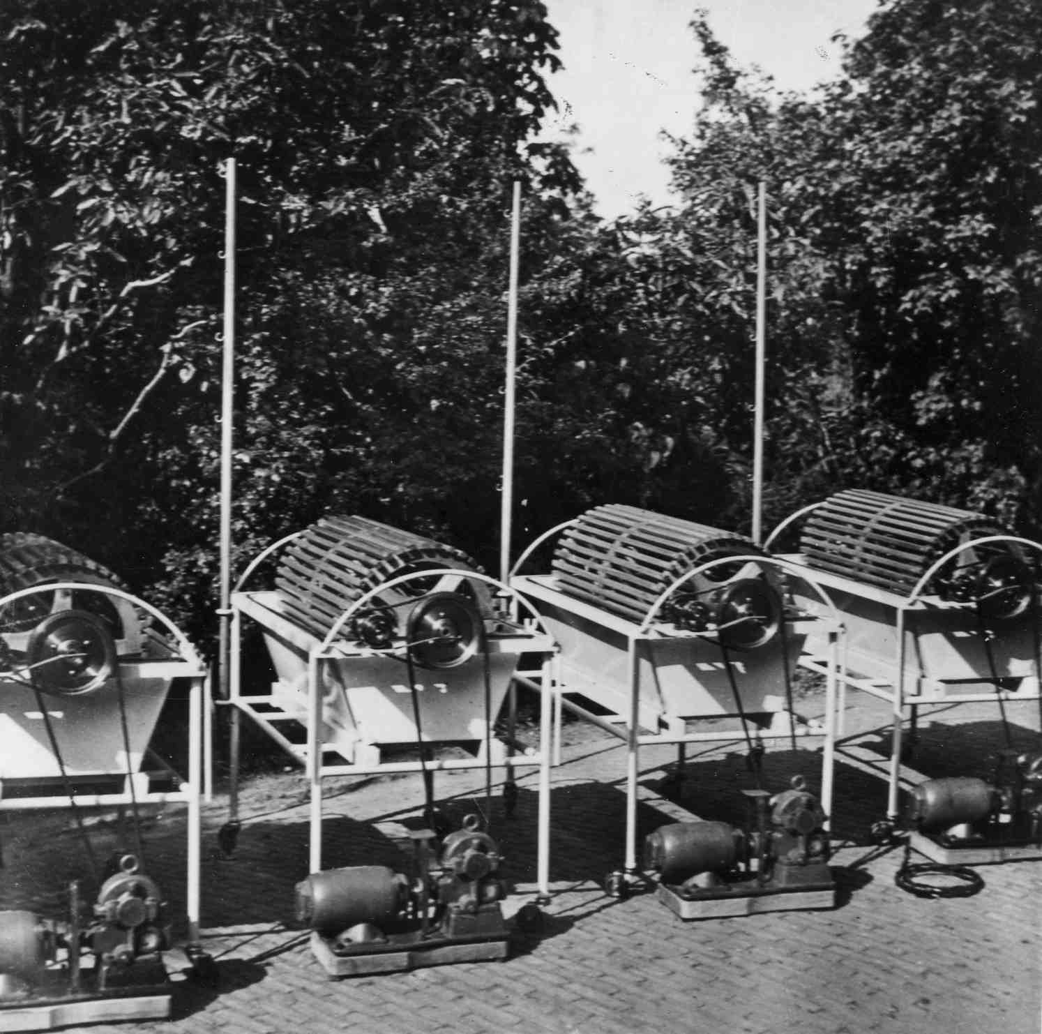 Four metal structures with cylindrical rollers and motors, set against a backdrop of trees, likely designed for a mechanical process.