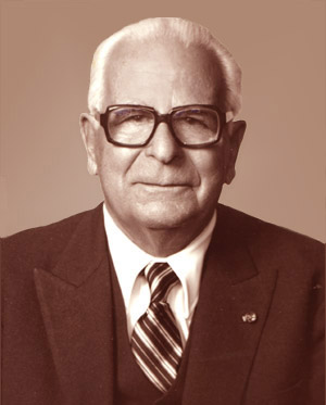  Portrait of Dr. Aziz Atiya, an older man with white hair, wearing large dark-rimmed glasses, a dark suit, a white shirt, and a striped tie. He has a calm expression and is facing the camera directly. The background is a soft brown gradient.
