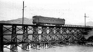 A vintage train crossing a wooden trestle bridge over water, surrounded by mountains and power lines, in black and white.