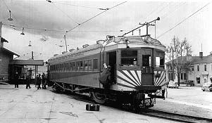 Black and white image of a vintage train at a station, with passengers boarding and a nearby building under a cloudy sky.