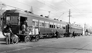 A vintage black-and-white photo of a train station, with workers unloading goods from a passenger train onto a horse-drawn cart.