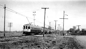 An early 20th-century electric train travels along tracks lined with utility poles in a rural setting.