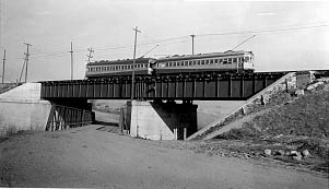 A vintage trolley crosses a steel bridge over a dirt road, with power lines and fields in the background.