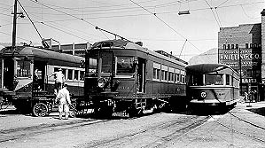 Vintage black-and-white photo of two electric streetcars at an intersection, with a man adjusting the tracks and buildings in the background.