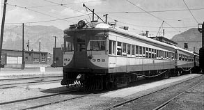 A vintage electric train, styled in cream and dark green, sits on tracks with mountains in the background and a railway station nearby.