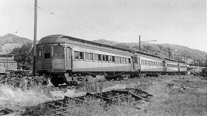 Black and white image of abandoned vintage train cars in a grassy area, set against a backdrop of rolling hills.