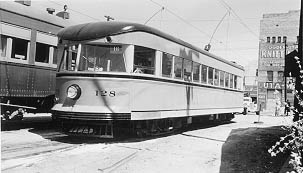 A vintage black-and-white photo of streetcar 128, showcasing its streamlined design, parked next to train cars and urban buildings.