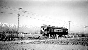 A vintage electric train car traveling along tracks with snow-capped mountains in the background, set in a serene rural landscape.