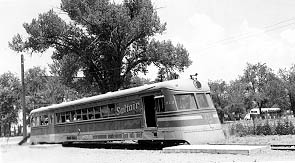 A vintage electric train car, streamlined and metallic, parked beside a large tree in a rural setting on a sunny day.