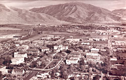 Aerial view of a historic University of Utah campus with buildings and tree-lined streets, set against a mountainous backdrop. Black and white photo.