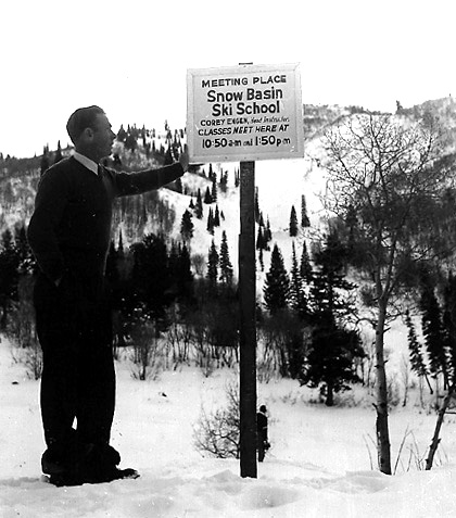 A man stands beside a sign at Snow Basin Ski School, detailing class times amidst a snowy landscape and evergreen trees.