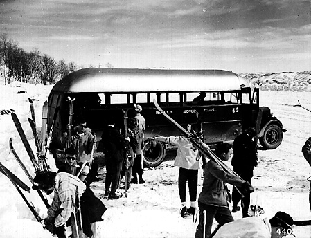 Skiers prepare for a snowy adventure near a vintage bus, surrounded by snow-covered hills and a clear winter sky.