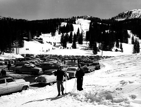 Two skiers stand in the snow, overlooking a crowded parking lot and ski slope surrounded by trees and mountains.