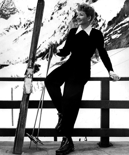 A stylish skier in a black outfit poses confidently at a mountain resort, with skis and poles by her side, snow-covered peaks in the background.