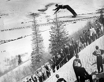 A vintage black and white photo of a ski jumper soaring through the air, while spectators watch from a snowy slope below.