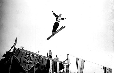 A black and white photo of a skier in mid-air, jumping off a ramp with arms outstretched, surrounded by banners and onlookers.