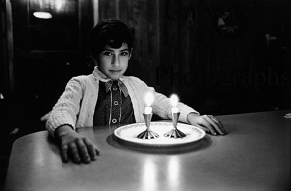 A child sits at a table in a dimly lit room, with two candles placed on a white plate in front of them, creating a warm atmosphere.