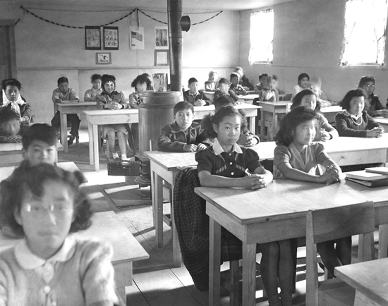 A historic black-and-white image of a school classroom filled with children seated at wooden desks, focused and attentive.